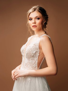 Lüks, parlak, dantelli gelinlik. Tüylü etekli ve derin dekolteli yaz kolsuz gelinliği. Çekici bayan gelin stüdyoda kahverengi arka planda poz veriyor. Doğal görünümlü makyaj ve saç stili..