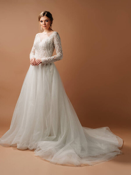 Luxury wedding dress. Fashionable bridal look. Ball gown with tender french lace and beads, long sleeves, lush tulle skirt. Beautiful young blonde bride in studio on brown background. Marriage concept
