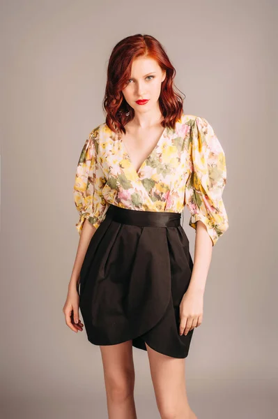 Female fashion. Redhead lady in elegant mini evening dress with long yellow balloon sleeves and black skirt on grey studio background. Young woman with red lips and natural hairdo. Holiday outfit.