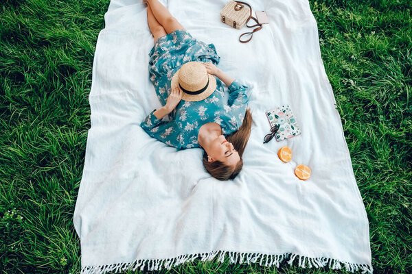 Beautiful woman in a colored dress and a straw hat lies on a white blanket on a background of green grass, women's accessories, summer time. Copy, empty space for text.