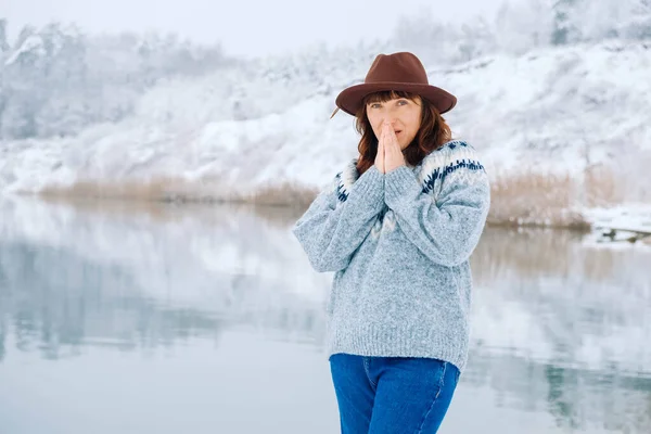 Женщина в коричневой шляпе и свитере согревает ладони на фоне зимнего леса и озера — стоковое фото