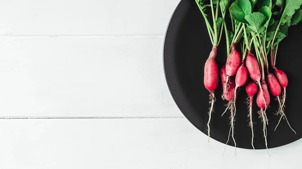 Bunch of radishes in a black plate on a white wooden background.