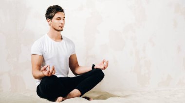 Young man sits in a meditative pose on sand against a wall background.