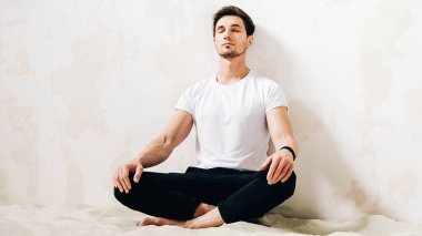 Young man sits in a meditative pose on sand against a wall background.