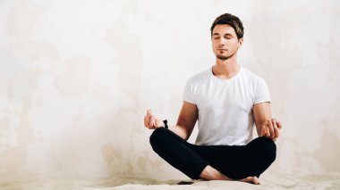 Young man sits in a meditative pose on sand against a wall background.