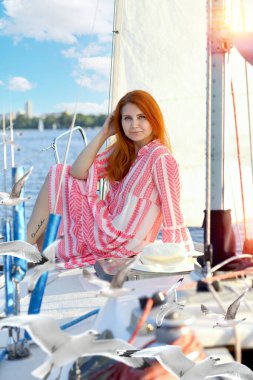 Pembe mini elbiseli mutlu kızıl saçlı bir kız martıların uçtuğu bir yatta oturuyor. Arka plan - şehir manzarası, beyaz yelkenler, mavi deniz, güneş ışınları. Seyahat kavramı