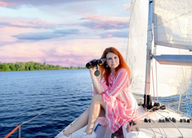 Beautiful red-haired girl in a pink mini dress sitting on a yacht with binoculars. The background is a stunning pink sunset, sky, white sails, sea. Summer. Travel concept. Copy space on the left