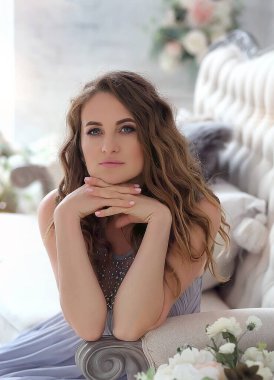 Güzel, genç, esmer bir kızın yakın portresi. Ellerini yüzünün yanına katlamış, lavanta rengi bir elbise giymiş. Modern iç mekan, arka planda çiçek dekoru. Moda ve güzellik konsepti