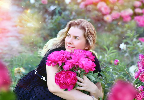 Beautiful and serene young woman hugging a bright bouquet of pink peonies. Background - a garden, rays of the sun, a peony field. Flowers and floristry concept