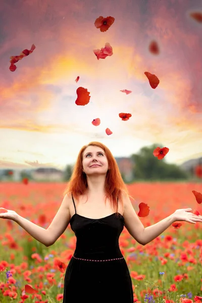 Beautiful young redhead woman in a black sundress in a poppy field where red petals fly. Flowers. Summer.  Fashion and beauty concept.