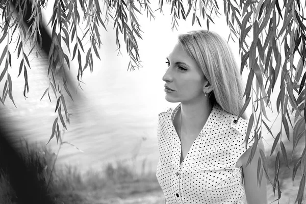 Black and white banner with a natural young blonde girl in a white dress sitting on the background of the river. Rest at nature. The concept of relaxation and meditation. Copy space on left