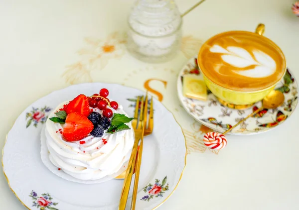 Meringue dessert decorated with strawberries, blackberries and red currants on a porcelain plate with cutlery and a cup of hot coffee. Morning concept.