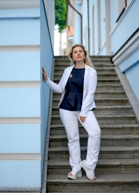 Beyaz takım elbiseli, kendinden emin ve dingin beyaz bir kadın merdivenlerde kameraya bakıyor. Mavi şehir binalarının arka planı. Moda ve güzellik. Sokak tarzı konsept.