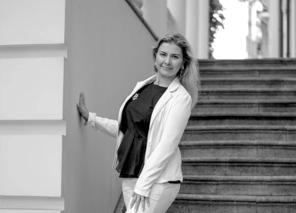 Black and white photo happy caucasian woman in white suit standing on the stairs smiling. Background of blue city buildings. Fashion and beauty. Urban style concept