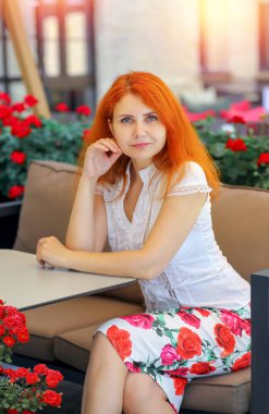 Beyaz bluzlu, kızıl saçlı ve şehvetli bir kızın portresi ve bir yaz kafesinde oturan gül desenli bir etek. Kameraya bakıyorum. Yazın şehir manzarası. Güzellik ve moda