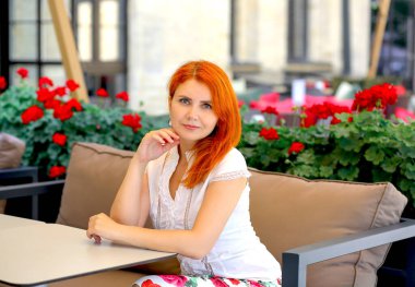 Kırmızı saçlı bir iplikle pankart ve beyaz bluzlu şehvetli bir kız ve bir yaz kafesinde oturan gül desenli bir etek. Kameraya bakıyorum. Yazın şehir manzarası. Dinlen ve rahatla.