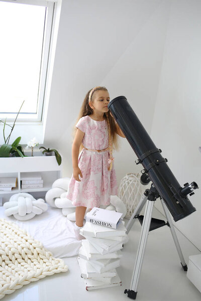 A cute little girl is looking at a telescope on an aluminum stargazing stand in a room on the attic floor. Equipment and technologies. Modern interior. Building a house concept
