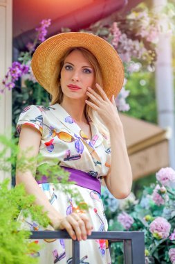 A stunning portrait of a blond girl in a wicker hat and elegant dress and against the backdrop of the city on a summer day. Fashion and beauty concept.