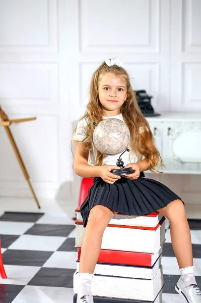 a cute Caucasian girl in a white blouse and a black pleated skirt sitting with a globe in her hands. Back to school. Education and childhood concept.