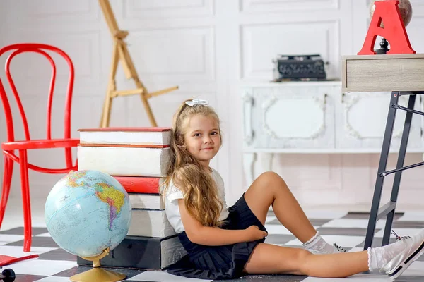 Cute aucasian girl in a white blouse and black pleated skirt sitting in a school interior. Back to school.Classroom with globe and school supplies.Education