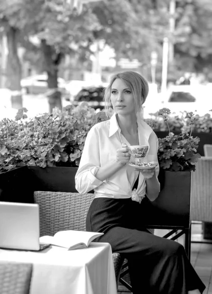 Black and white photo with lady sitting in a summer cafe with a cup of coffee in a white shirt. Coffee break. The background is an urban landscape. business concept