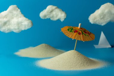 Oyuncak güneş şemsiyesi, kum, bulutlar, mavi arka planda kağıt tekne. Fotokopi alanı olan yaratıcı plaj tatili konsepti. Dinlenme, deniz ve tatil kavramı.