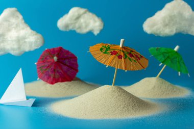 Oyuncak güneş şemsiyesi, kum, bulutlar, mavi arka planda kağıt tekne. Fotokopi alanı olan yaratıcı plaj tatili konsepti. Dinlenme, deniz ve tatil kavramı.