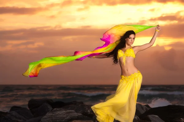 A beautiful bellydancer poses with a flowing yellow silk veil and standing on a rocky bank in front of the Atlantic Ocean — Stock Photo, Image