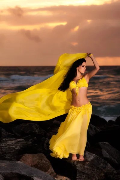 A beautiful bellydancer poses with a flowing yellow silk veil and standing on a rocky bank in front of the Atlantic Ocean — Stock Photo, Image