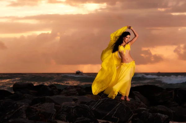 A beautiful bellydancer poses with a flowing yellow silk veil and standing on a rocky bank in front of the Atlantic Ocean — Stock Photo, Image