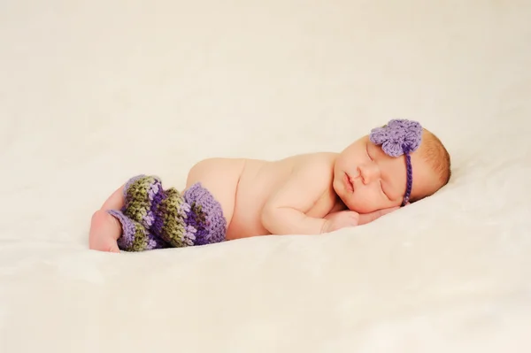 Newborn baby girl wearing purple and green crocheted legwarmers and headband and sleeping on a cream colored blanket. — Stok fotoğraf