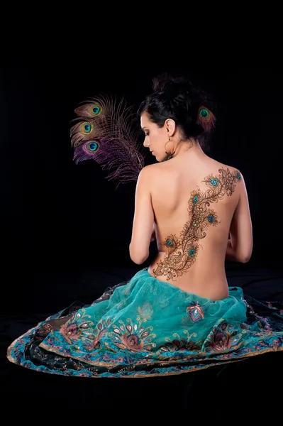 Mujer sentada en el suelo y sosteniendo tres plumas de pavo real. Ella tiene un diseño de henna de plumas de pavo real en su espalda . — Foto de Stock