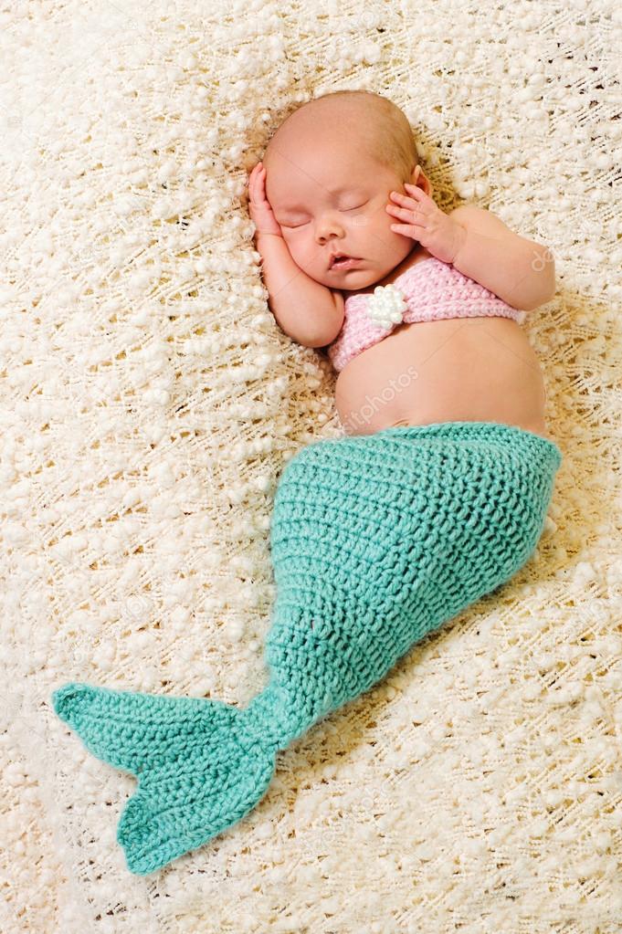 Newborn baby girl wearing a crocheted turquoise and pink mermaid costume, sleeping on a cream colored blanket.
