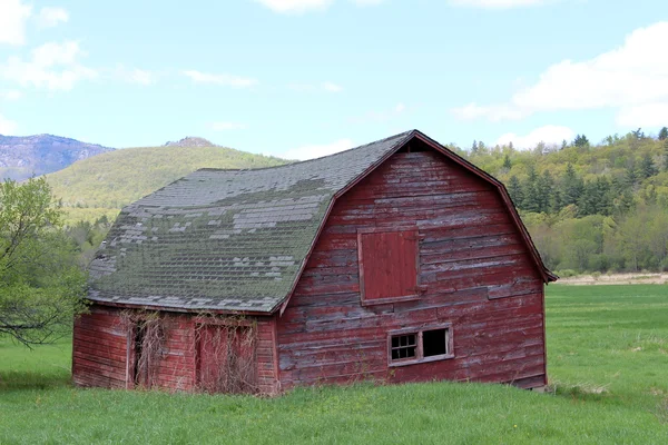 Old abandoned red barn out in rural countryside with mountain range and blue skies as backdrop — Stock Photo, Image