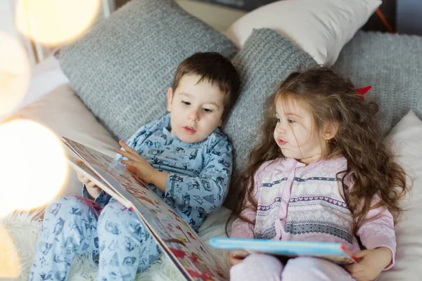 Toddler boy and girl in pyjamas lying on the bed and read books near Christmas tree in light Christmas interior. Cute Christmas kids