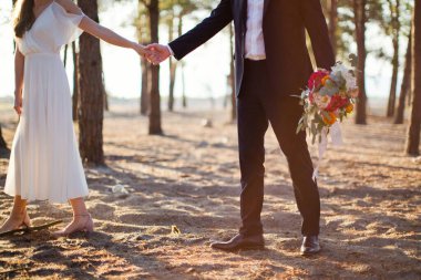 Gelin ve damat el ele tutuşup gün batımında deniz kenarında yürürler.