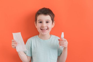Okul öncesi beyaz çocuk beyaz tıbbi maske ve turuncu arka planda dezenfektan taşıyor. Coronavirus önleme kavramı