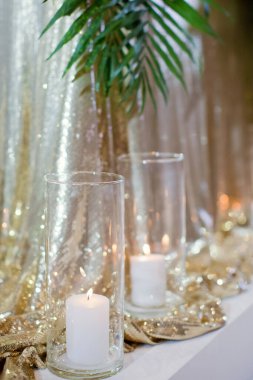 Altın pullu masa örtüsüyle düğün masasının yanında palmiye yaprağı olan cam vazolarda mum yakmak.