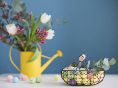 Çiçekli ve yumurtalı Paskalya süslemeleri. Sarı sulama kabındaki güzel çiçekler ve klasik metal sepetteki renkli yumurtalar.