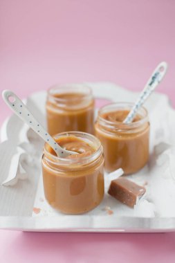 Karamel soslu kavanozlar ve kağıda sarılmış karamel şekerler pembe arka planda beyaz bir ahşap tepside yatıyorlar.