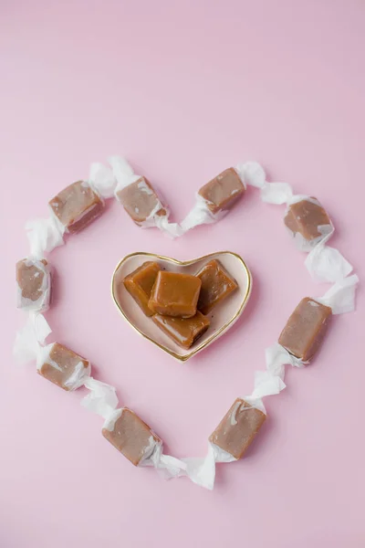 Heart shaped caramel candy fudge in paper wrapping on a pink background with copy space