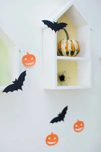 Halloween decorations and pumpkins. Black bats and orange pumpkins from paper on a white wall. House shaped shelves with pumpkins and spiders