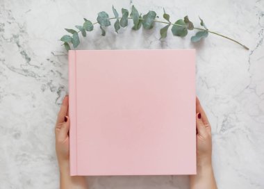 Pink cover book with copy space on a marble background and eucalyptus twig. Woman holding pink book album
