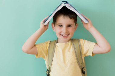 Caucasian preschool boy with school backpack holds book on a light green background with copy space