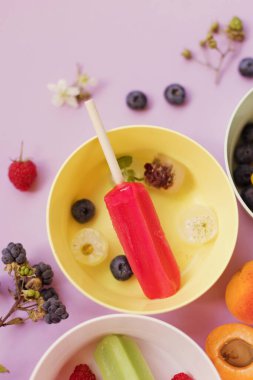 Bright colorful popsicles in colorful bowls with berries and ice cubes on a light violet background. Tasty ice pops close up