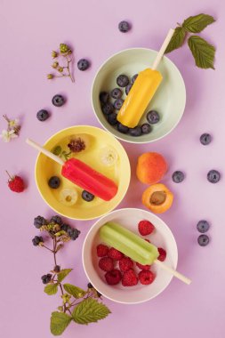 Bright colorful popsicles in colorful bowls with berries and ice cubes on a light violet background. Tasty ice pops close up