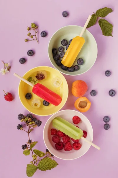 Bright colorful popsicles in colorful bowls with berries and ice cubes on a light violet background. Tasty ice pops close up