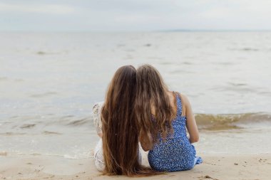 Two caucasian women with dark long hair sitting on the sand and looking on sea. Friend an girlfriends concept