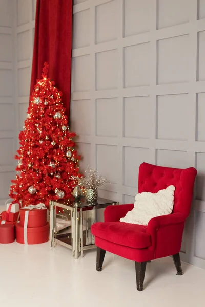 Interior with red Christmas tree, gift boxes, glass table and red vintage armchair near grey wall