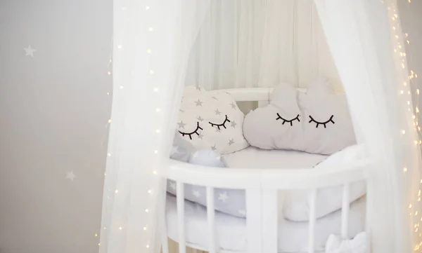 White childs bed crib with canopy and lights near white wall with copy space. Cute infant baby crib with pillows. Light kids interior